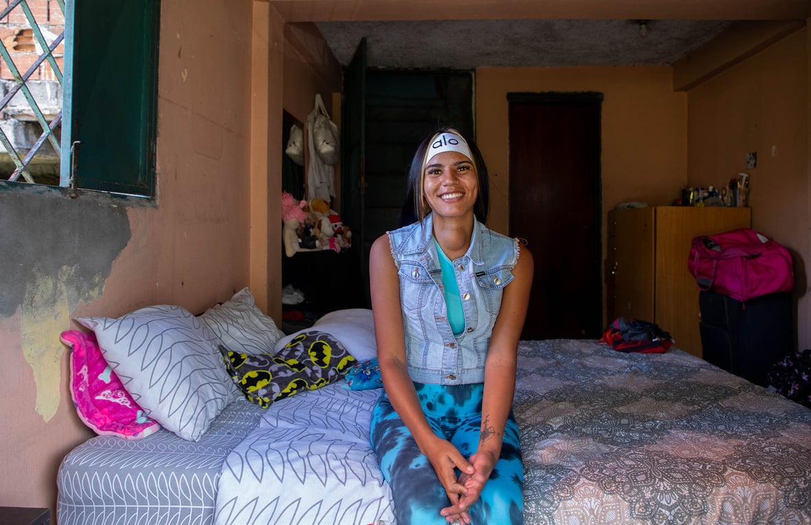 Una mujer joven posa sonriendo sentada al borde de una cama tendida en una habitación colorida.