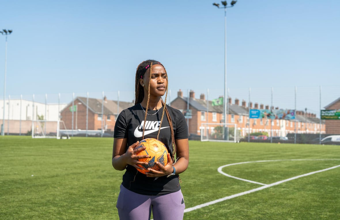 Una joven sostiene un balón de fútbol en una cancha de fútbol con hileras de casas al fondo
