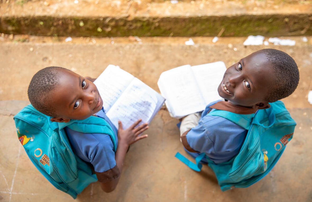 Dos niñas de 7 y 8 años, procedentes de la República Centroafricana, sonríen a la cámara desde donde están sentadas al aire libre, con mochilas a juego en las que se lee 'La educación no puede esperar' y sosteniendo libros abiertos