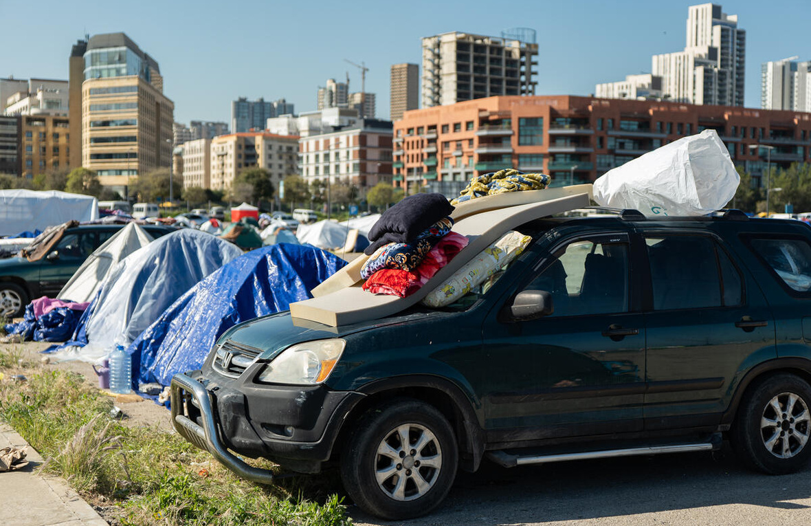 Tiendas de campaña y un vehículo con colchones y almohadas en un lote de estacionamiento cerca de algunos edificios.