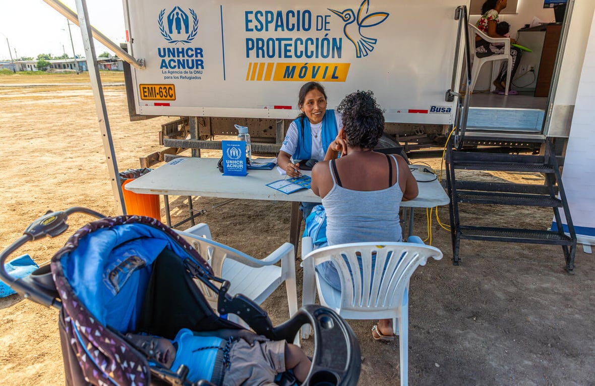 Una mujer con uniforme de ACNUR, camiseta blanca y chaleco azul, habla con una mujer que se encuentra de espaldas sentada en una silla plástica blanca