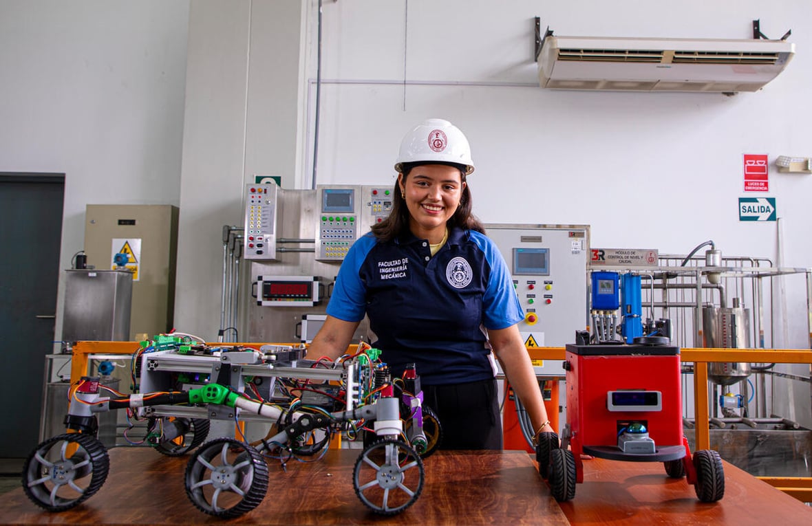 Yuliveth posa para una foto en un laboratorio rodeada dispositivos y mostrando en la mesa delante de ella varios modelos de pequeños robots.