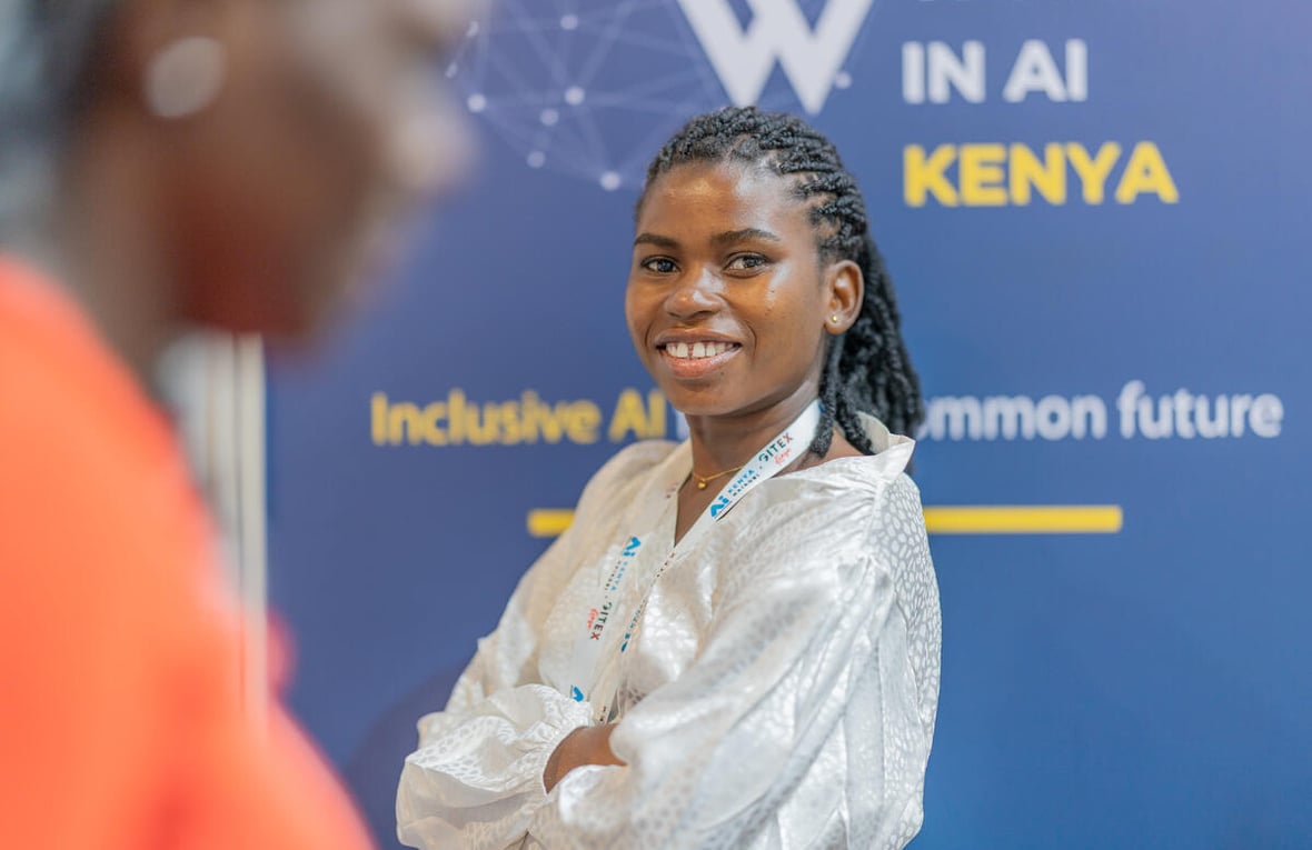 Una joven sonriente, con los brazos cruzados, posa delante del cartel de una conferencia sobre las mujeres en el ámbito de la IA en Kenia