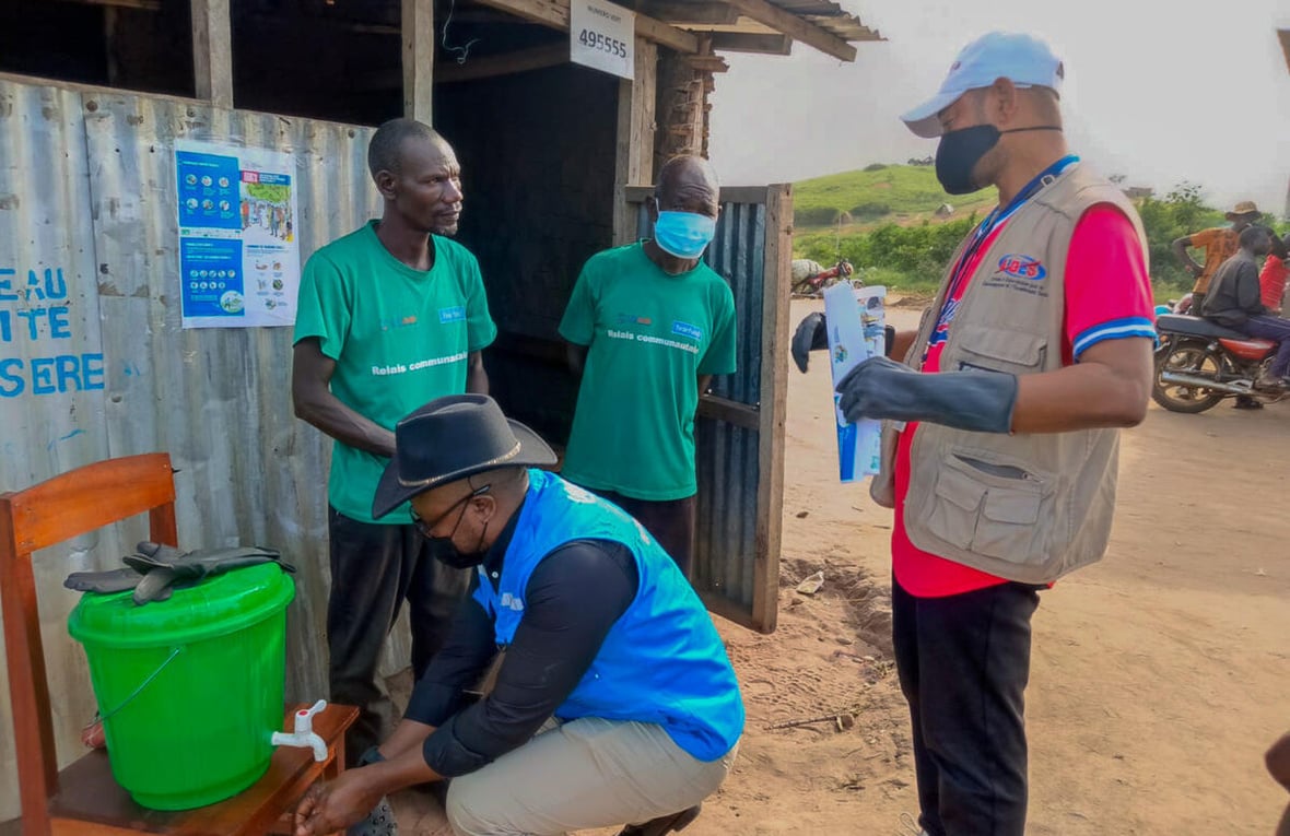 Cuatro hombres, tres de ellos con cubrebocas, afuera de un alojamiento esperan para recoger agua de un contenedor