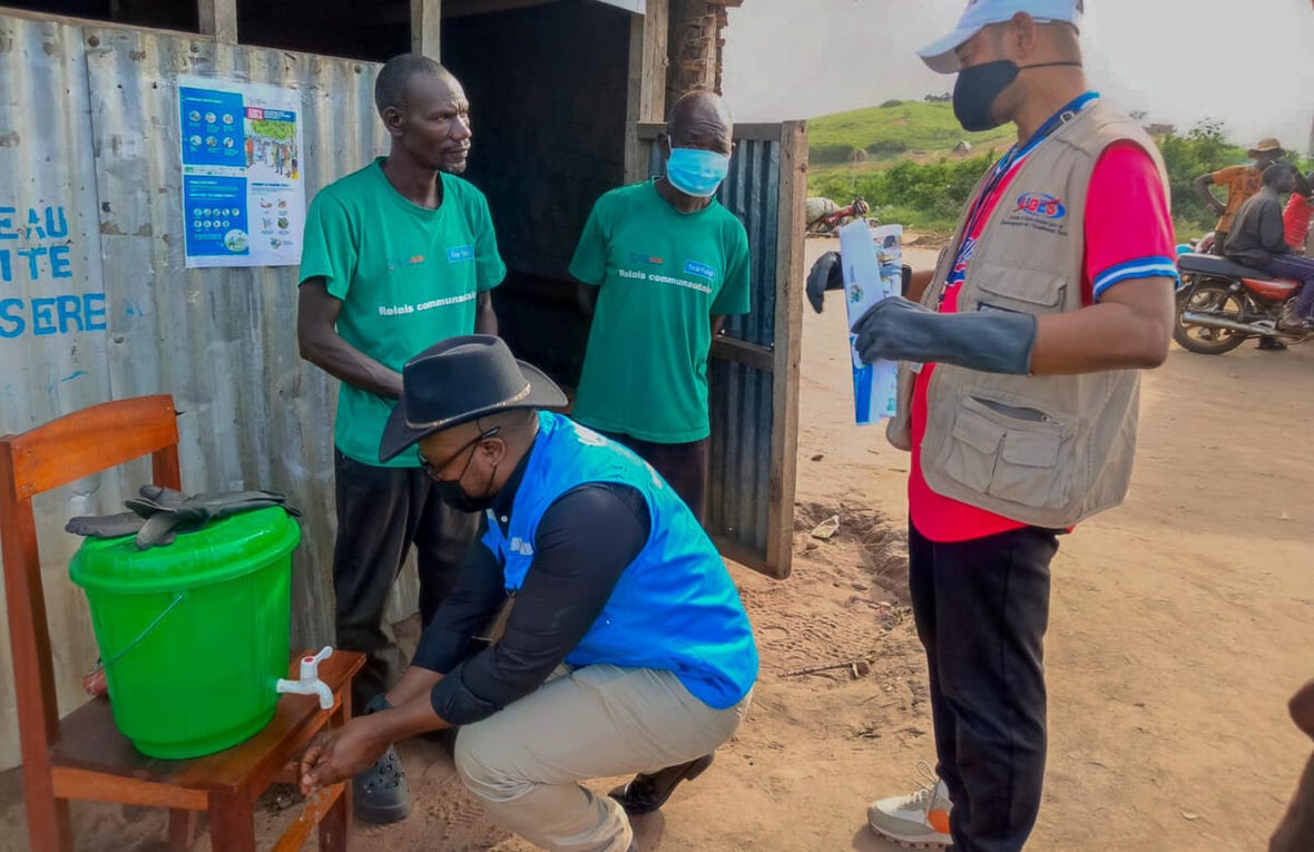 Cuatro hombres, tres de ellos con cubrebocas, afuera de un alojamiento esperan para recoger agua de un contenedor