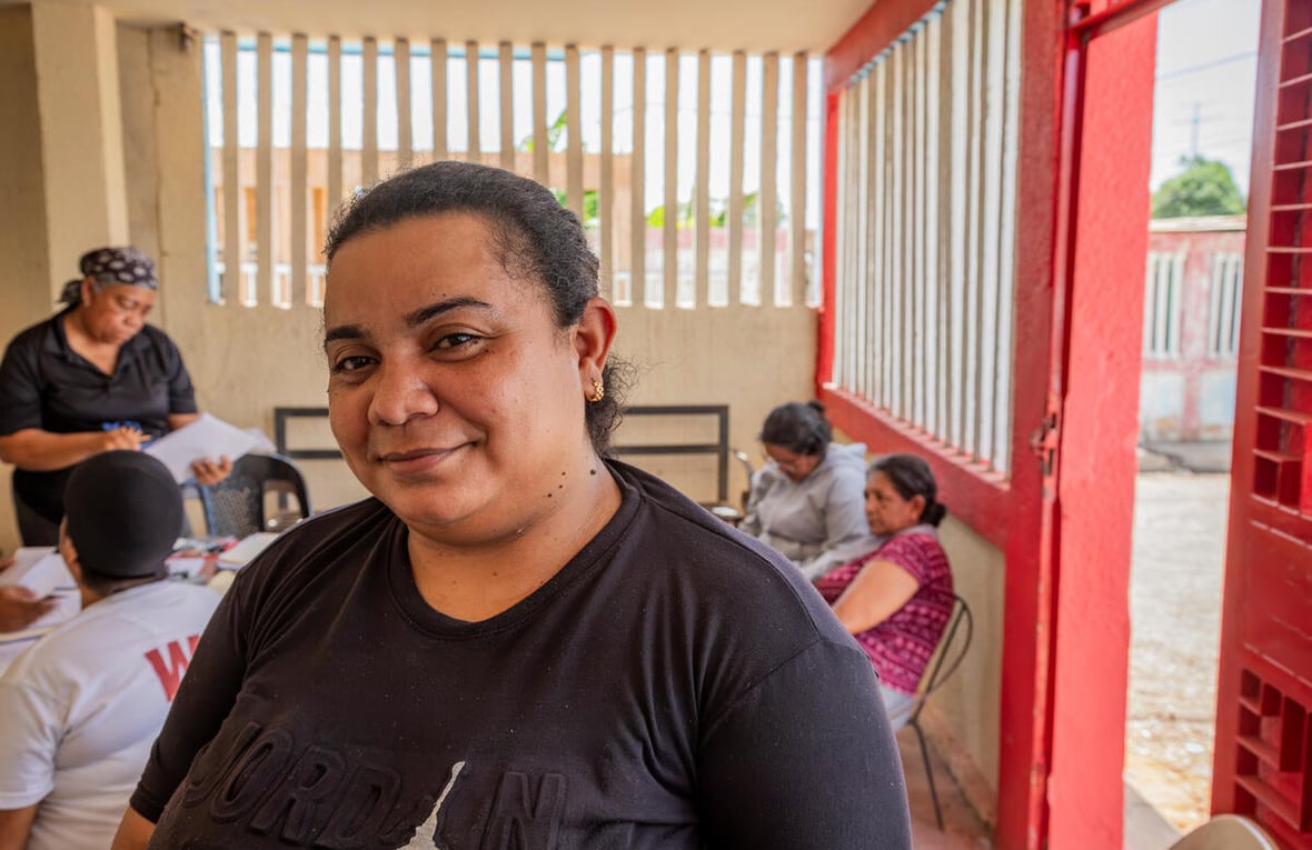 Una mujer posa para la cámara en un salón donde se encuentran varias personas realizando trámites.