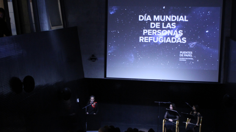 En un auditorio oscuro se lee en una pantalla con letras blancas: DÍA MUNDIAL DE LAS PERSONAS REFUGIADAS