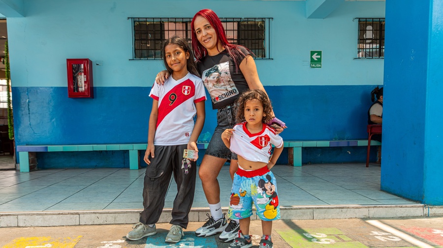 Una mujer de cabello rojo, posa con su hija y su hijo pequeño en un patio de suelo colorido. Al fondo una pared en dos tonos de azul