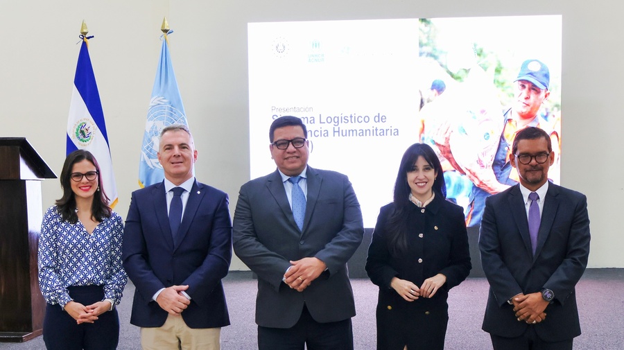 Tres hombres y dos mujeres con vestimenta formal, de pie frente a un foro y dos banderas al fondo