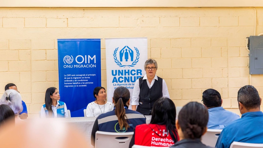 Una mujer hablando desde una mesa de ponencia en un evento frente a un público sentado en una sala, carteles con los logos de ACNUR y OIM están al fondo.