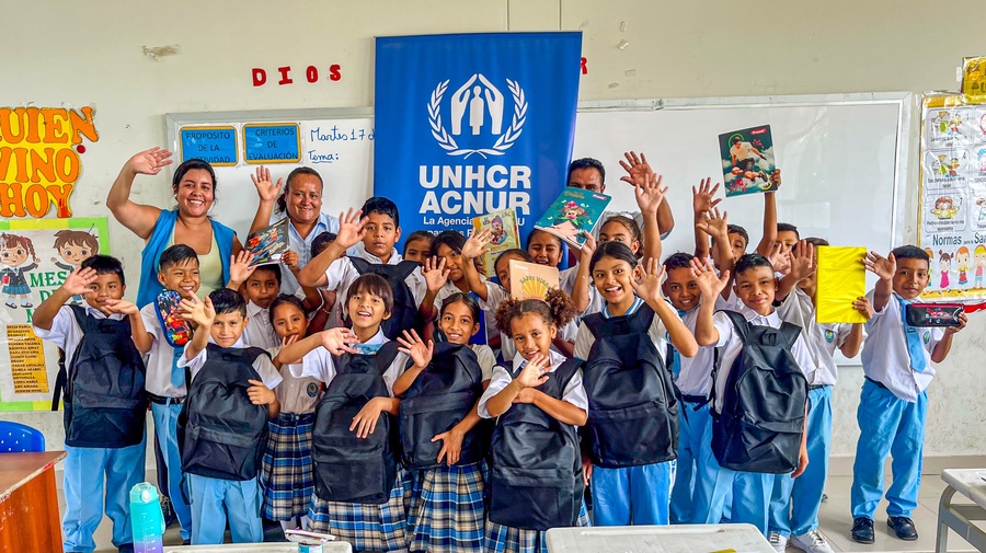 Una veintena de niñas y niños y tres profesores saludan a la cámara en un salón de clases