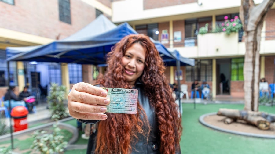 Una mujer de cabello largo rizado sostiene en primer plano con su mano derecha un carné de la República del Perú