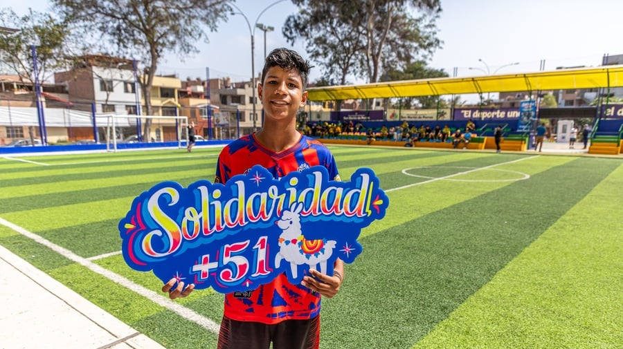 Un joven que viste uniforme de fútbol rojo, sostiene un letrero colorido en el que se lee SOLIDARIDAD +51 en una cancha de fútbol