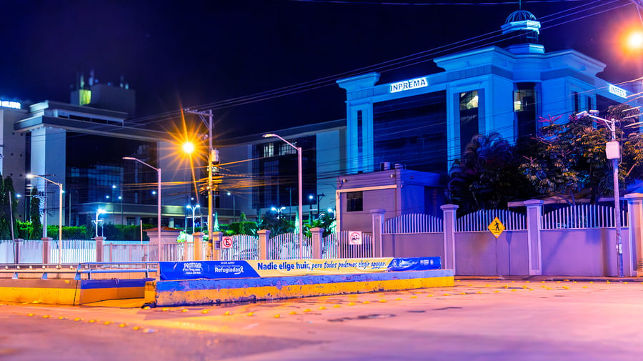 Un edificio iluminado de azul. En una barda de lee "nadie elige huir, pero todos podemos elegir apoyar"