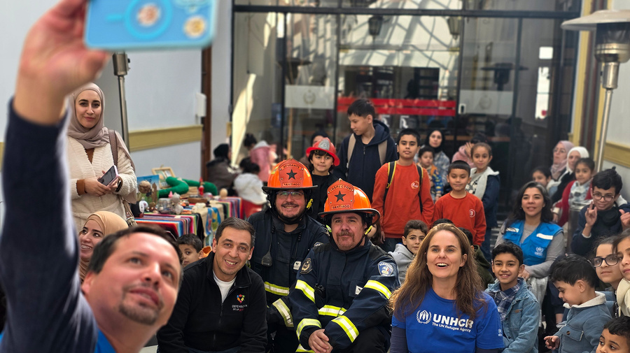 Al menos una decena de niñas y niños posan para la foto junto a ocho adultos, entre ellos dos con vestimenta de ACNUR en color azul y otros dos hombres con uniforme y casco de bombero