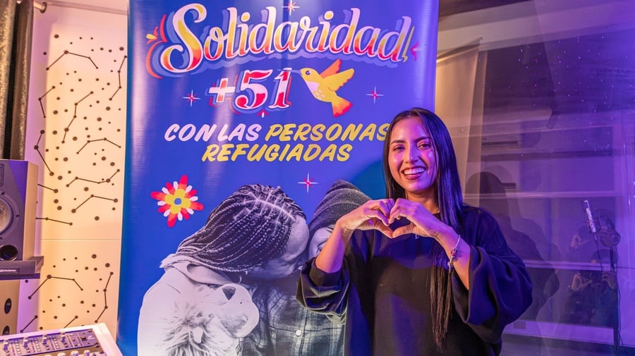 Una mujer de cabello largo oscuro hace un corazón con sus manos mientras sonríe y muestra su dentadura. Al dondo, un letrero azul con letras amarillas donde se lee SOLIDARIDAD +51 CON LAS PERSONAS REFUGIADAS