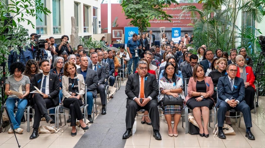 Decenas de personas sentadas en filas en un jardín. Al fondo se observan dos letreros de ACNUR en color azul