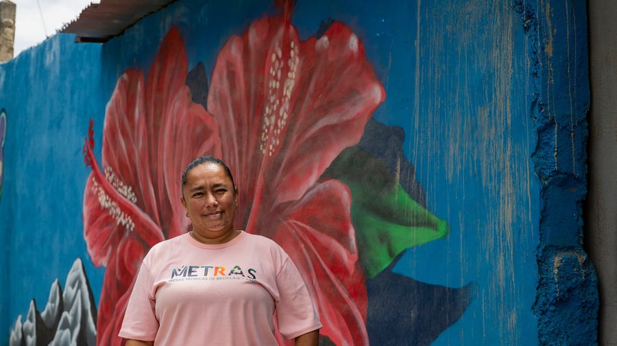 Una mujer con camiseta rosa, de pie frente a una pared azul que tiene pintada una flor roja grande con hojas verdes