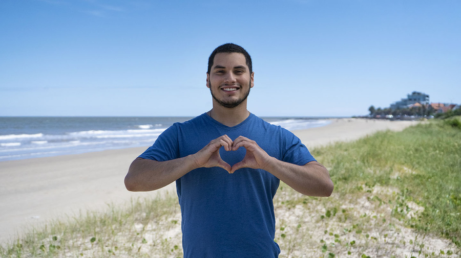Un joven posa para la cámara en una playa haciendo el gesto del corazón con las manos.