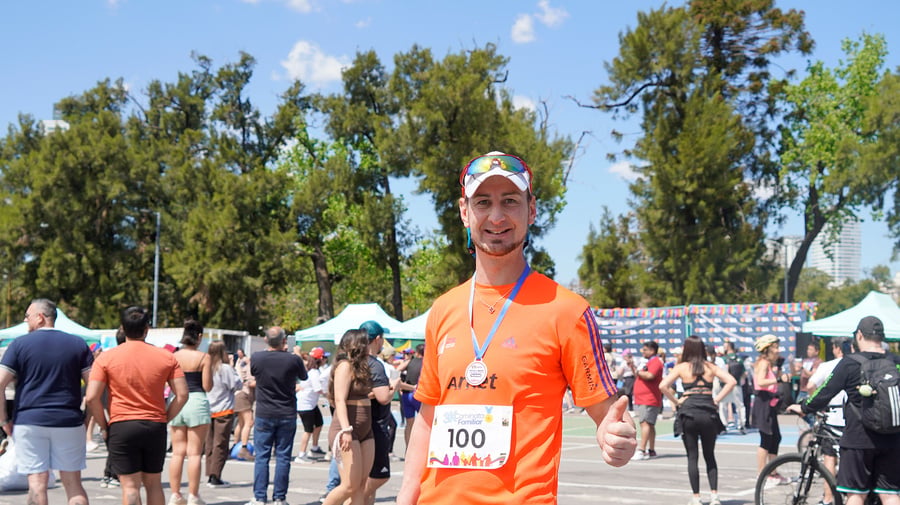 Un hombre con camiseta naranja y shorts negros, porta una medalla colgada al cuello mientras levanta su pulgar izquierdo al sonreir para la foto