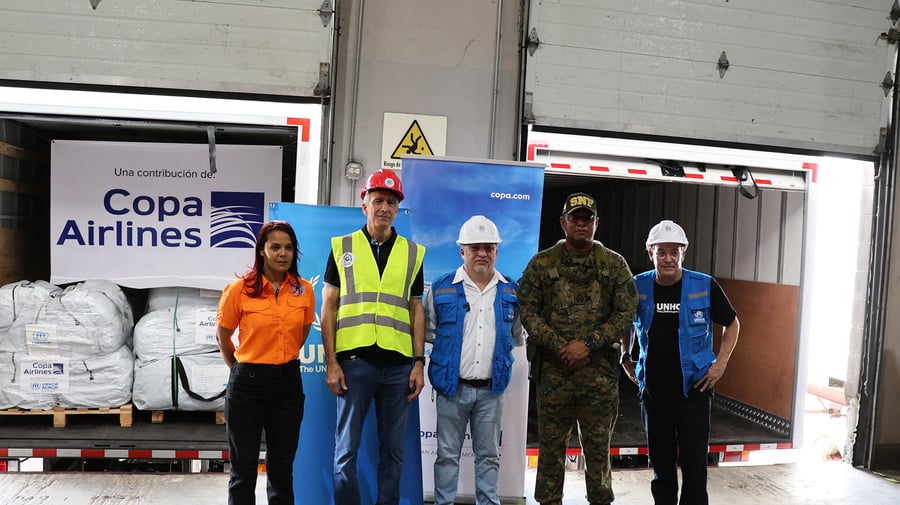 Una mujer que viste pantalon negro y camisa naranja, seguida de 4 hombres que tienen vestimenta de trabajo, tres de ellos usan casco