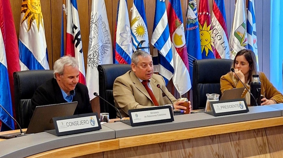 Dos hombres y una mujer sentados en una mesa semicircular con banderas de diferentes países al fondo