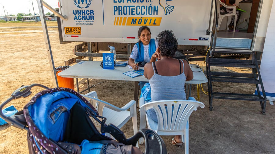 Una mujer con uniforme de ACNUR, camiseta blanca y chaleco azul, habla con una mujer que se encuentra de espaldas sentada en una silla plástica blanca