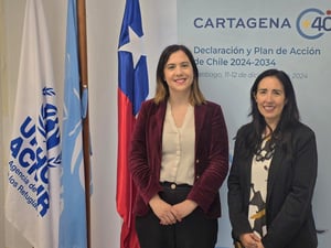 Dos mujeres posan para la cámara al lado de banderas de Chile, Naciones Unidas y ACNUR. A sus espaldas un rótulo mencional el Plan de Chile y la campaña Cartagena+40.