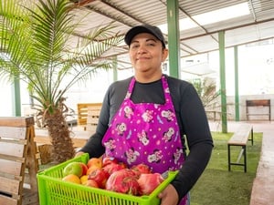 Una mujer sonríe a la cámara cargando una caja de frutas coloridas.