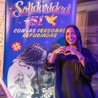 Una mujer de cabello largo oscuro hace un corazón con sus manos mientras sonríe y muestra su dentadura. Al dondo, un letrero azul con letras amarillas donde se lee SOLIDARIDAD +51 CON LAS PERSONAS REFUGIADAS