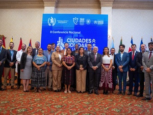 Un grupo de 14 hombres con traje, y 7 mujeres con vestimenta formal, posan para la foto frente a banderas de diferentes países. Al fondo, en letrero azul con letras blancas se lee: CIUDADES SOLIDARIAS