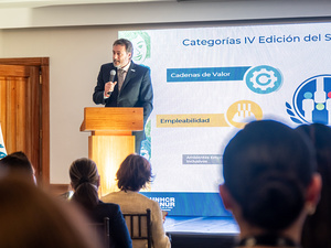 Un hombre de traje hablando desde un atril en un evento en un espacio cerrado con varios asistentes.