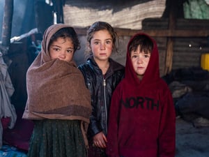 Dos niñas y un niño están de pie frente a su alojamiento, vestidos con ropa de abrigo