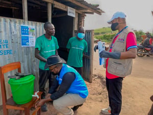 Cuatro hombres, tres de ellos con cubrebocas, afuera de un alojamiento esperan para recoger agua de un contenedor