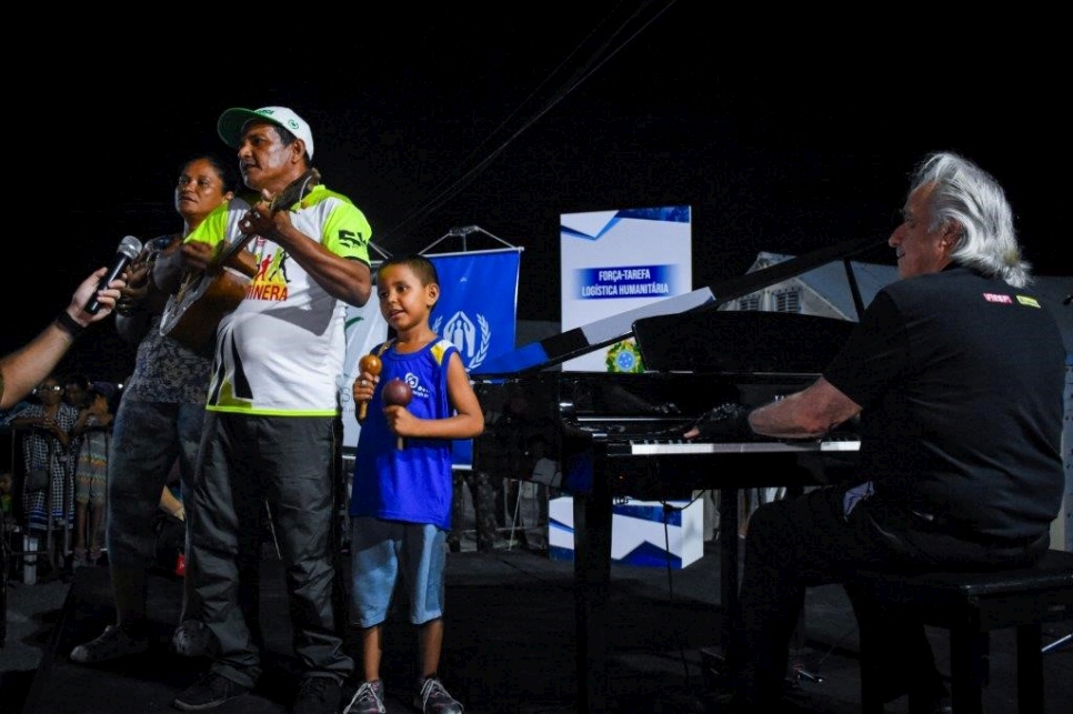 Guillermo, Katty y el pequeño Arnel actuando en el escenario junto al director de orquesta brasileño João Carlos Martins, mientras la población del albergue canta con ellos.