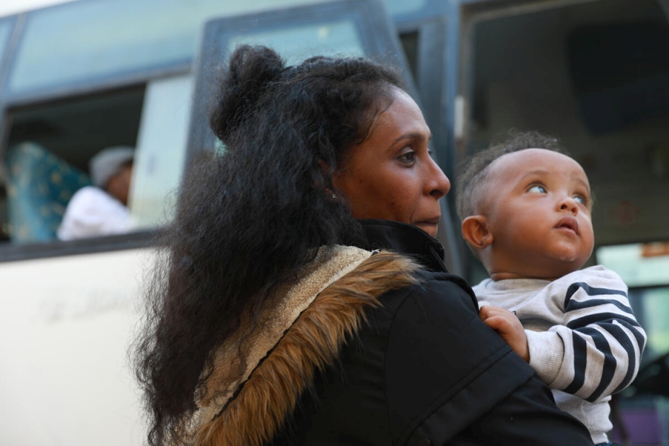 Solicitante de asilo lleva a su bebé en brazos mientras se prepara para abordar un autobús con dirección al aeropuerto, donde tomarán un vuelo para salir de Libia.