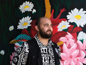 De perfil, un hombre con vestimenta blanco con negro frente a un mural de flores blancas y rosas
