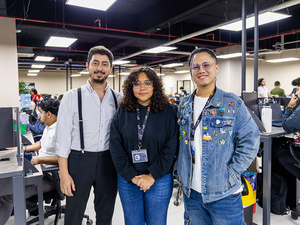 Dos hombres y una mujer posan juntos para una foto en una oficina abierta con escritorios, computadoras y personal trabajando.