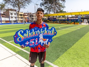 Un joven que viste uniforme de fútbol rojo, sostiene un letrero colorido en el que se lee SOLIDARIDAD +51 en una cancha de fútbol