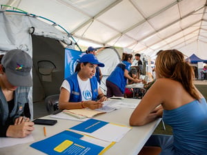 Detrás de una mesa, una trabajadora de ACNUR con vestimenta azul de la organización, atiende a una mujer que está de espaldas del otro lado de la mesa