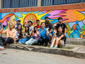 Jóvenes posan para una foto de grupo sentados en una acera frente a un mural colorido.