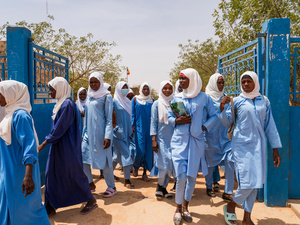 Un grupo de jovencitas sudanesas atraviesa una reja color azul; cada una de ellas lleva la cabeza cubierta con un manto color blanco y porta un atuendo azul claro.