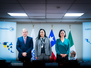 Un hombre y dos mujeres posan para una foto formal en un salón. A sus espaldas respecivamente las banderas de ACNUR, de Chile y de México. A los lados dos afiches con el logo del proceso de Cartagena+40.