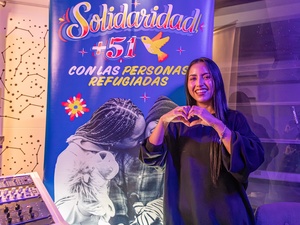 Una mujer de cabello largo oscuro hace un corazón con sus manos mientras sonríe y muestra su dentadura. Al dondo, un letrero azul con letras amarillas donde se lee SOLIDARIDAD +51 CON LAS PERSONAS REFUGIADAS