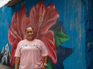 Una mujer con camiseta rosa, de pie frente a una pared azul que tiene pintada una flor roja grande con hojas verdes
