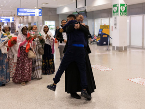 Dos hombres vestidos de negro se abrazan fuertemente en un aeropuerto, mientras que un grupo de mujeres permanece cerca, sonriendo y aplaudiendo