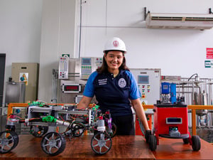 Yuliveth posa para una foto en un laboratorio rodeada dispositivos y mostrando en la mesa delante de ella varios modelos de pequeños robots.
