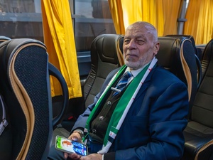 Un hombre mayor de saco y corbata sentado en un bus sujeta un celular en sus manos y sonríe mirando hacia la cámara.