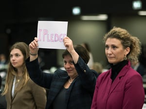 Tres mujeres forman una hilera. Las que están a los extremos son rubias; la que está en el centro, de cabello corto y oscuro, sostiene frente a sí un cartel que dice "Me comprometo".