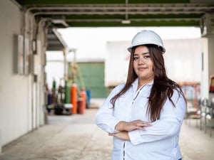 Una joven salvadoreña sonríe a la cámara; lleva puesta una blusa blanca y un casco de seguridad del mismo color. Tiene los brazos cruzados.